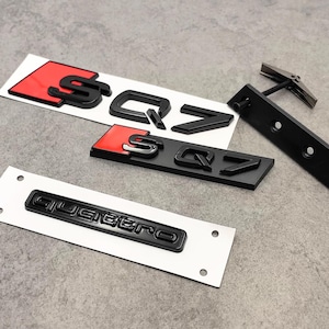 Peut inclure: Plusieurs emblèmes de voiture noirs et rouges, dont les badges "SQ7" et "Quattro", sont présentés sur une surface grise. Les badges "SQ7" comportent un fond rouge. Un support de montage noir est également visible.