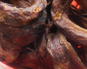 May include: A close-up shot of a red bowl filled with dried, smoked fish. The fish is a deep brown color with a slightly charred appearance. The bowl sits on a white surface, with a green background.