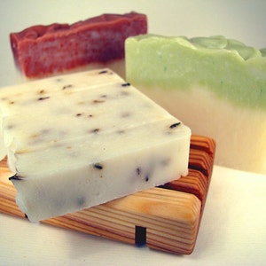 May include: Three bars of handmade soap on a wooden soap dish. The soap bars are in shades of red, green, and white. The soap dish is made of light brown wood.