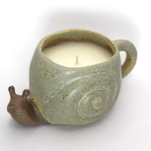 May include: A ceramic candle shaped like a snail, with a light green body and a brown head. The candle is filled with a white wax and has a single wick. The snail's shell features a white spiral design and a handle.