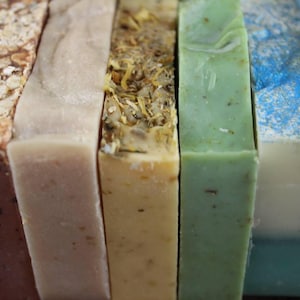May include: A close-up of a row of six different colored bars of soap. The soaps are arranged in a row from left to right, with the colors being brown, beige, yellow, green, blue, and light blue. The soaps are all rectangular in shape and have a smooth surface.