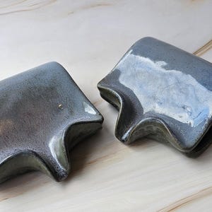 May include: Two ceramic soap dishes with a unique, angular design. The dishes are a muted blue-gray color with a glossy finish, and they have a curved, wave-like edge. The soap dishes are displayed on a light-colored, marbled surface.