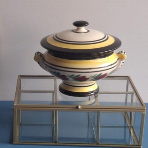 Op de afbeelding: Een houten bord met de tekst "ENTREZ, C'est ouvert" in het wit, met pijlen en een hart. Een decoratieve keramische kom met een deksel, met gele en zwarte strepen, staat op een glazen en metalen vitrine. De kom heeft bloemmotieven.