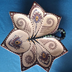 Peut inclure: Un plat en céramique en forme d'étoile avec un motif floral. Le plat est de couleur crème avec des accents marron, violets et jaunes. Il a une poignée vert foncé et est posé sur un fond bleu. Plat de service décoratif.