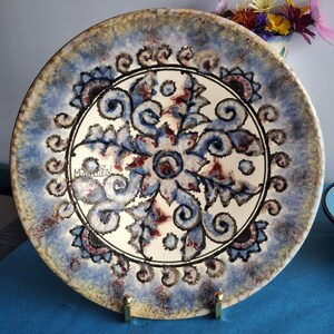 Op de afbeelding: Een ronde keramische plaat met een handgeschilderd bloemenontwerp. De plaat heeft een centraal bloemmotief in blauw, rood en wit, omgeven door een blauwe en bruine rand. De plaat wordt gepresenteerd op een goudkleurige standaard.