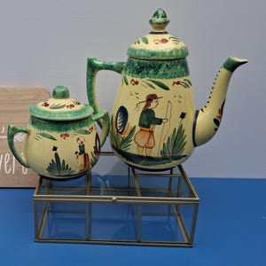 May include: A vintage ceramic teapot and creamer set with a yellow and green color scheme. The set features hand-painted figures and floral designs. A wooden sign with French text is also present.
