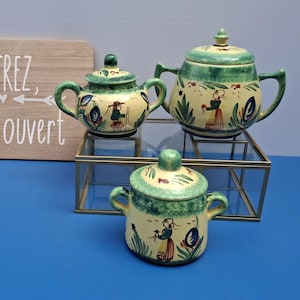 May include: Three ceramic teapots with lids, in varying sizes, are displayed on a glass and metal shelf. The teapots are painted with a yellow base, green accents, and figures. A wooden sign with the text "ENTREZ, C'est ouvert" is in the background.