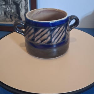 Może przedstawiać: Ceramiczna miska z dwoma bocznymi uchwytami, posiadająca głęboką niebieską glazurowaną górną część z pasem ukośnych beżowych pasków oraz ciemnobrązową glazurowaną dolną część. Wnętrze miski jest w kolorze jasnego beżu, a krawędź jest gładka.