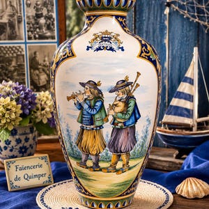 Può includere: Un vaso in ceramica con una scena dipinta a mano di due figure in abiti tradizionali che suonano il flauto e la cornamusa. Il recipiente color crema presenta bordi decorativi blu e gialli, uno stemma con la scritta BRETAGNE e un bordo svasato.