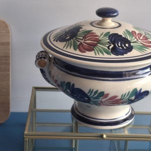 May include: A ceramic tureen with a lid, featuring a floral design in blue, red, and green on a cream background. The tureen has a pedestal base and handles. It sits on a glass display case.