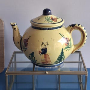 Peut inclure: Une théière en céramique jaune avec un couvercle, ornée de motifs floraux et figuratifs peints à la main. La théière a une anse et un bord bleu foncé, et est présentée sur une boîte en verre et en laiton. C'est un article de cuisine décoratif.