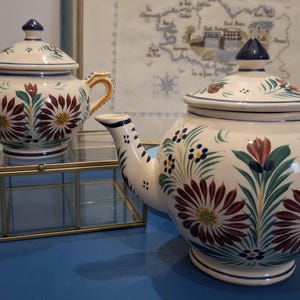 Może przedstawiać: Dwa ceramiczne zestawy do herbaty z kwiatowymi wzorami. Dzbanek do herbaty i cukiernica są białe z bordowymi, zielonymi i żółtymi akcentami. Dzbanek ma dzióbek i uchwyt, a cukiernica dwa uchwyty. Oba mają pokrywki z niebieskimi gałkami.