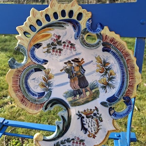 Puede incluir: Un plato decorativo de cerámica con borde festoneado, que presenta una escena pintada a mano de dos figuras tocando instrumentos. El plato tiene un fondo blanco con detalles en azul, amarillo y marrón. El plato está sobre una superficie azul.