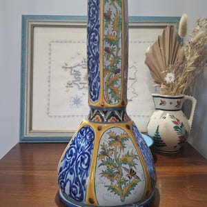 Puede incluir: Un jarrón decorativo de cerámica, alto y rectangular, con base acampanada. Presenta intrincados diseños florales y geométricos en azul, amarillo, blanco y un toque de naranja. El jarrón mide aproximadamente 61 cm de altura.