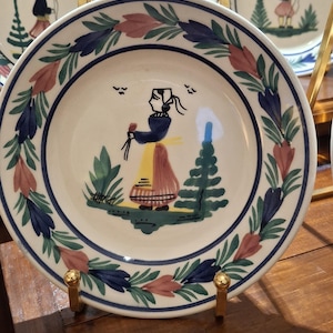 Puede incluir: Plato de cerámica color crema con un diseño pintado a mano. El plato presenta un borde de hojas azules, rosas y verdes. La imagen central representa una figura con vestimenta tradicional sosteniendo flores cerca de un árbol estilizado. Se ven platos adicionales.