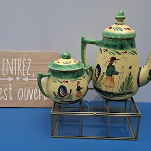 Può includere: Un set vintage di teiera e zuccheriera in ceramica con disegni floreali e figurativi verdi e gialli. La teiera è su un supporto di metallo. Un cartello di legno con testo francese è sullo sfondo.