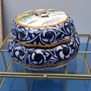 Puede incluir: Una caja de baratijas redonda de cerámica con tapa. La caja está decorada con patrones florales azules y blancos y un borde amarillo. La tapa presenta una escena pintada con figuras. La caja está sobre una vitrina de vidrio y metal.