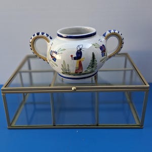 May include: A white ceramic creamer with two handles, decorated with a painted scene of a person holding a flower. The handles have blue and white patterns. The creamer sits on a glass and metal display case, with a blue background.