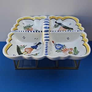 Può includere: Un piatto da portata in ceramica bianca con quattro scomparti e un manico. Il piatto presenta disegni dipinti a mano di sciatori, uccelli e motivi floreali in blu, rosso, giallo e verde. Poggia su un supporto in metallo dorato.
