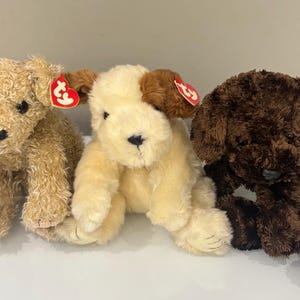 Peut inclure: Trois peluches en forme de chiens, dans des tons de marron et de crème. Chaque chien a une texture et une couleur de fourrure différentes, avec des yeux et des nez noirs. Chaque chien a une étiquette rouge en forme de cœur attachée à son oreille.