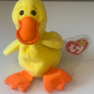 May include: A yellow plush duck toy with an orange beak and feet. The duck has black eyes and small wings. A red heart-shaped tag is attached to the duck.