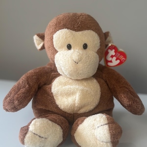 以下が含まれることがあります： 茶色の毛皮とクリーム色の顔、胸、足を持つぬいぐるみのおもちゃの猿。ぬいぐるみには、耳に赤いハート型のタグが付いています。猿は直立しています。