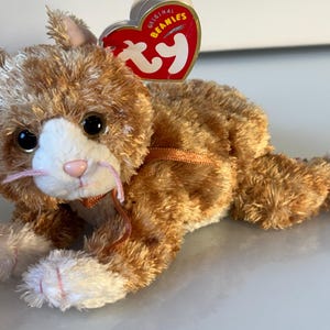 Op de afbeelding: Een pluche kattenspeeltje met oranje en witte vacht, zwarte ogen en een roze neus. Het heeft lange roze snorharen en een dun oranje lint om de nek. Een hartvormig rood en goudkleurig label met de tekst Original Beanies en ty is bevestigd.