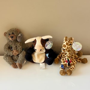 May include: Three plush toys: a brown teddy bear, a cow with black and white patches, and a giraffe with brown spots. Each toy has a tag attached. The toys are sitting on a white surface.
