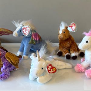 Puede incluir: Una colección de seis animales de peluche, incluyendo dragones, unicornios y caballos. Los juguetes son de varios colores, como marrón, morado, blanco y azul, con detalles como alas y crines. Cada juguete tiene una etiqueta.