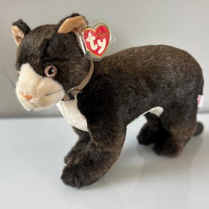 May include: A plush cat toy with brown and white fur, tan ears, and a red heart-shaped tag. The cat has a tan collar and whiskers. The toy is in a standing position.