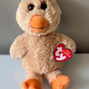May include: A plush duck toy with soft, light brown fur, orange feet and beak, and large, glittery eyes. It has a red heart-shaped tag that reads "Attic Treasures".