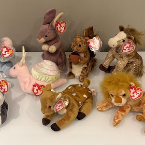 Peut inclure: Collection de peluches d'animaux, dont un éléphant, un lapin, un écureuil, un cheval, un lion, un cerf, un escargot et un husky. Chaque peluche a une étiquette en forme de cœur. Les jouets sont de différentes couleurs et tailles, exposés sur une surface blanche.
