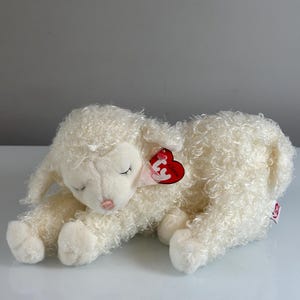 Puede incluir: Un peluche de cordero color crema con pelo rizado, tumbado. Tiene los ojos cerrados, una nariz rosa y una cinta rosa. Una etiqueta roja en forma de corazón está sujeta a su oreja.