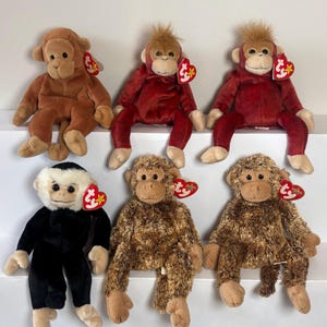 May include: Six plush monkey toys in various shades of brown and red. Each monkey has a distinct facial expression and a red heart-shaped tag. The monkeys are sitting on a white surface.