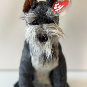 Op de afbeelding: Een pluche Schnauzer hondenspeelgoed met grijs en wit haar, bruine oren en een zwarte neus. Het speelgoed heeft een lange, witte baard en een rood hartvormig label aan zijn oor. Het speelgoed zit rechtop.