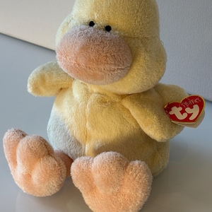 May include: A plush yellow duck toy with soft fur, a rounded body, and light orange feet and beak. The duck features small black eyes and a red heart-shaped tag attached to its wing that reads Ty Pluffies in white lettering.