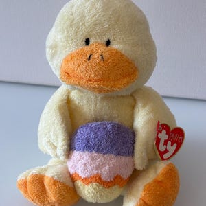 May include: A plush yellow duckling toy with a soft, fuzzy texture, featuring an orange beak and orange feet. The duckling is holding a small, rounded object with horizontal stripes in purple, light pink, and orange. A red heart-shaped Ty tag is attached.