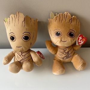 Pode incluir: Duas figuras de pelúcia Groot marrons com traços faciais detalhados e linhas corporais costuradas. Uma figura tem os braços ao lado, a outra tem os braços levantados. Cada uma tem uma etiqueta em forma de coração Ty.