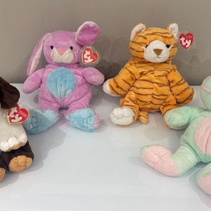 Puede incluir: Cuatro peluches: un perro, un conejo, un tigre y una rana. El perro es marrón, blanco y negro. El conejo es rosa y azul. El tigre es naranja y marrón. La rana es verde y rosa.