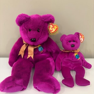May include: Two plush purple teddy bears of different sizes. Both bears have black eyes, a gold ribbon, and a small earth emblem. Each bear has a Ty heart-shaped tag attached.