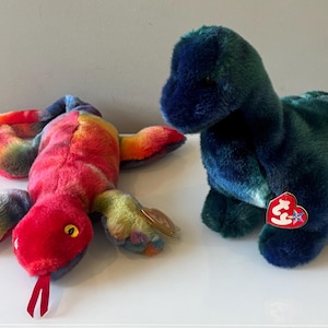 Puede incluir: Dos peluches: una serpiente colorida con cabeza roja y un dinosaurio azul y verde. La serpiente es de color arcoíris con una lengua roja. El dinosaurio es azul oscuro y verde con una etiqueta roja en forma de corazón.