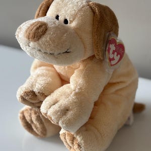 Op de afbeelding: Een pluche hond in tinten beige en bruin. De hond heeft bruine oren, neus en poten, met een zachte, donzige textuur. Een rood hartvormig label is aan het oor bevestigd. De hond zit rechtop.