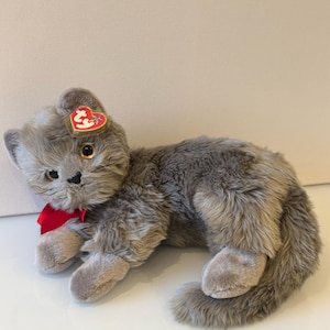 May include: A plush gray cat toy with a red bow and a heart-shaped tag. The cat is lying down with its paws extended. The toy has a soft, furry texture and detailed facial features.