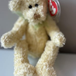 May include: A small, cream-colored teddy bear with a green ribbon around its neck. The bear has black eyes and a brown nose. A red tag with white text is attached to its ear. The bear is sitting.