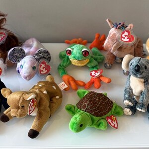 May include: A selection of plush animal toys, including a panda, deer, frog, koala, and bear. Each toy has a tag attached. The toys are in various colours, such as brown, green, and black.