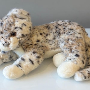 May include: A soft toy snow leopard in a lying pose. The plush animal features a cream-coloured body with black spots and white paws. It has a furry texture and detailed facial features, including whiskers and brown eyes.