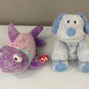 Puede incluir: Dos juguetes de peluche: un pez colorido y un perro azul. El pez es principalmente morado con detalles rosas y verdes, y el perro es azul claro con orejas y patas azul oscuro. Ambos juguetes tienen ojos grandes y expresivos.