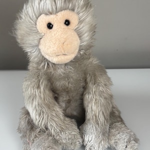 May include: A soft toy monkey with grey fur and a pale tan face. The monkey has black eyes and is in a sitting position. The fur is long and shaggy, giving the toy a soft appearance. The toy is designed to resemble a monkey.