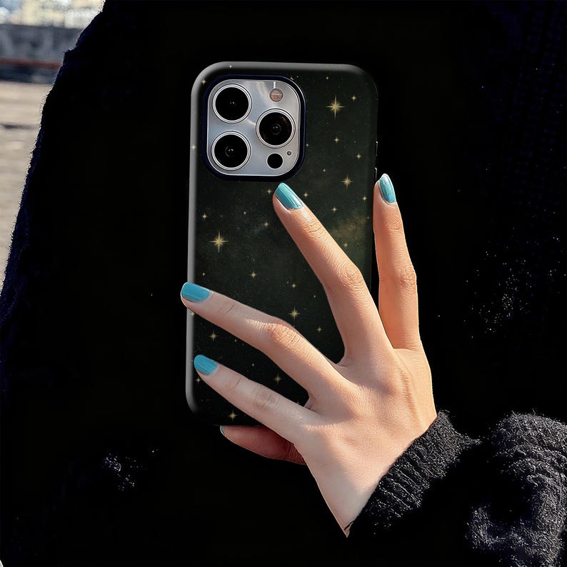 May include: Black phone case with a starry night design. The case has a dark background with small gold stars scattered across it. The phone case is held by a hand with blue nail polish.
