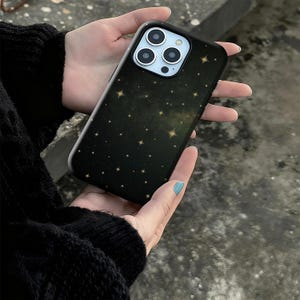May include: Black phone case with a starry night design. The case features a dark background with numerous gold stars scattered across the surface. The phone case has a black border and a camera cutout.